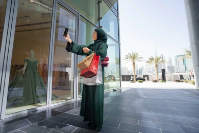 الاستفادة من العروض الموسمية والتخفيضات، استخدام التكنولوجيا في الحياة اليومية، إمرأة سعودية تقوم بالتسوق وشراء المستلزمات والهدايا عبر الانترنت،سيدة عربية خليجية اماراتية ترتدي الحجاب والعباءة تمسك بيدها الهاتف المحمول وأكياس التسوق، التقاط صور السيلفي التذكارية