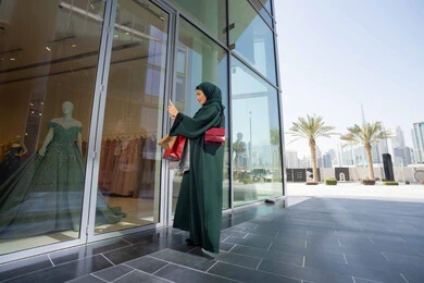 Buying new items from the mall, enjoying shopping and getting offers, taking advantage of seasonal discounts, an Emirati Gulf Arab woman wearing a hijab and abaya holding colorful shopping bags and taking pictures of a dress with her mobile phone with gestures of pleasure and joy