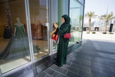 Enjoying shopping and getting offers, buying new items from the mall, taking advantage of seasonal discounts, an Emirati Gulf Arab woman wearing a hijab and abaya holding colorful shopping bags and taking pictures of a dress with her mobile phone with gestures of pleasure and joy