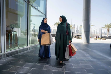 Enjoying shopping in malls and shopping centers, choosing supplies and gifts with friends, two Arab Gulf Emirati women wearing the abaya and hijab holding paper bags shopping with gestures of cheerfulness and enthusiasm