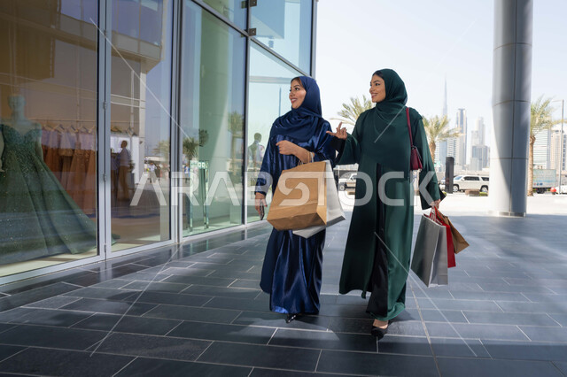 Taking advantage of the days of discounts and exciting offers, buying necessities from the mall, choosing supplies and gifts with friends, two Arab Gulf Emirati women wearing the abaya and hijab holding paper bags shopping with gestures of cheerfulness and enthusiasm