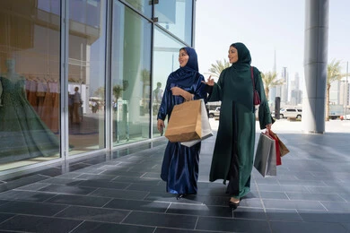 Taking advantage of the days of discounts and exciting offers, buying necessities from the mall, choosing supplies and gifts with friends, two Arab Gulf Emirati women wearing the abaya and hijab holding paper bags shopping with gestures of cheerfulness and enthusiasm