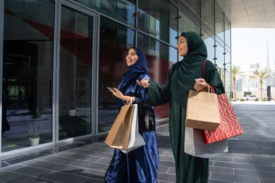 الشراء في أيام التنزيلات والتخفيضات، الاستمتاع بالشراء والتسوق ، متعة التسوق مع الاصدقاء، سيدتان عربيتان خليجيتان اماراتيان ترتديان العباءه و الحجاب تحملان حقائب التسوق , الجمعه البيضاء