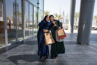 Women's shopping obsession, the beauty of markets in the Emirates, choosing supplies and gifts with friends, two Arab Gulf Emirati women wearing the abaya and hijab holding paper bags, pointing and looking into a distant place with gestures of cheerfulness and enthusiasm