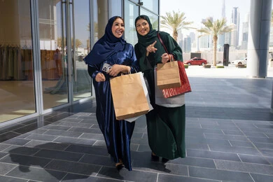 The positive impact of shopping and buying on women, choosing supplies and gifts with friends, two Arab Gulf Emirati women wearing the abaya and hijab holding paper bags, pointing and looking into the distance with gestures of cheerfulness and enthusiasm