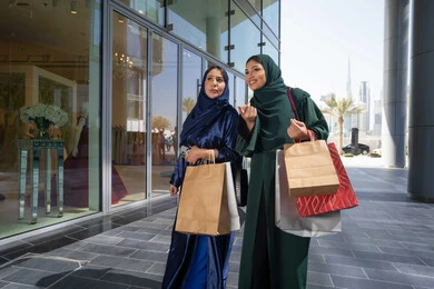 اختيار المستلزمات والهدايا برفقة الأصدقاء ،امراتان عربيتان خليجيتان اماراتيان يرتديان العباءه و الحجاب تمسكان بالأكياس الورقية تشيران وتنظران إلى مكان بعيد بإيماءات البشاشة والحماس