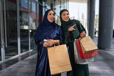 الاستمتاع بالشراء والتبضع، إقتناء المستلزمات من المول ،امراتان عربيتان خليجيتان اماراتيان يرتديان العباءه و الحجاب تمسكان بالأكياس الورقية تشيران وتنظران إلى مكان بعيد بإيماءات البشاشة والحماس