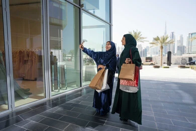 الاستفادة من العروض الموسمية والتخفيضات، متعة مشاركة الأصدقاء في اختيار الأشياء والمستلزمات من المولات التجارية، الأثر الإيجابي للتسوق على النساء، شابتان عربيتان خليجيتان اماراتيان مبتسمتان ترتديان الحجاب و العباءه تحملان الأكياس الورقية تقومان بالتبضع في المول، التقاط  صور سلفي عن طريق الجوال