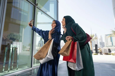 متعة مشاركة الأصدقاء في اختيار الأشياء والمستلزمات من المولات التجارية، مواكبة آخر صيحات الموضة، الأثر الإيجابي للتسوق على النساء، شابتان عربيتان خليجيتان اماراتيان مبتسمتان ترتديان الحجاب و العباءه تحملان الأكياس الورقية تقومان بالتبضع في المول، التقاط صور سلفي عن طريق الجوال