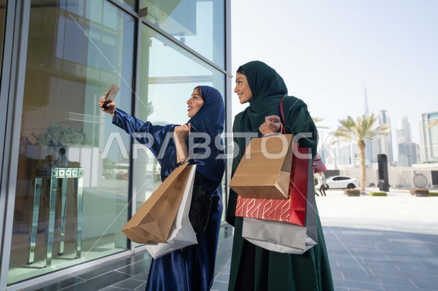 ايماءات السعادة و السرور، متعة مشاركة الأصدقاء في اختيار الأشياء والمستلزمات من المولات التجارية، الأثر الإيجابي للتسوق على النساء، شابتان عربيتان خليجيتان اماراتيان مبتسمتان ترتديان الحجاب و العباءه تحملان الأكياس الورقية تقومان بالتبضع في المول، التقاط صور سلفي عن طريق الجوال