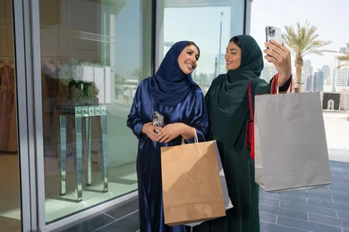استخدام الجوال في التسوق، متعة مشاركة الأصدقاء في اختيار الأشياء والمستلزمات من المولات التجارية، الأثر الإيجابي للتسوق على النساء، شابتان عربيتان خليجيتان اماراتيان مبتسمتان ترتديان الحجاب و العباءه تحملان الأكياس الورقية يقومان بالتبضع في المول، التقاط صور سلفي تذكارية