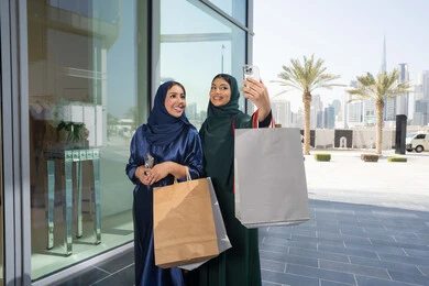 Benefiting from Black Friday discounts, the joy of sharing with friends in choosing things and supplies from shopping malls, keeping up with the latest fashion trends, two smiling Emirati Gulf Arab girls wearing hijab and abaya carrying paper bags shopping in the mall, taking selfies with their mobile phones