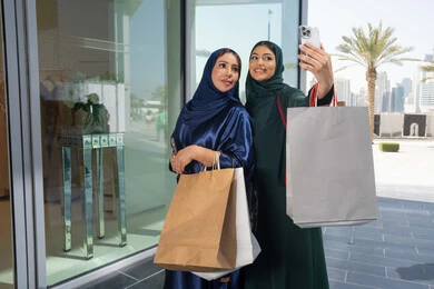 الأثر الإيجابي للتسوق على النساء، متعة مشاركة الأصدقاء في اختيار الأشياء والمستلزمات من المولات التجارية، مواكبة آخر صيحات الموضة، شابتان عربيتان خليجيتان اماراتيان مبتسمتان ترتديان الحجاب و العباءه تحملان الأكياس الورقية تقومان بالتبضع في المول، التقاط صور سلفي عن طريق الجوال
