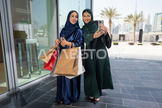 Keeping up with the latest fashion trends, the joy of sharing with friends the selection of things and supplies from shopping malls, the positive impact of shopping on women, two smiling Emirati Gulf Arab girls wearing hijab and abaya carrying paper bags shopping in the mall, taking selfies with their mobile phones