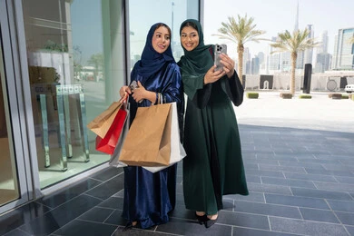 مواكبة آخر صيحات الموضة، متعة مشاركة الأصدقاء في اختيار الأشياء والمستلزمات من المولات التجارية، الأثر الإيجابي للتسوق على النساء، شابتان عربيتان خليجيتان اماراتيان مبتسمتان ترتديان الحجاب و العباءه تحملان الأكياس الورقية تقومان بالتبضع في المول، التقاط صور سلفي عن طريق الجوال