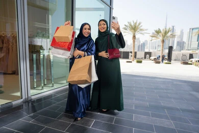 The joy of shopping with friends, buying gifts and supplies, taking selfies with the mobile phone, taking advantage of discounts and sales, two Emirati Arab women wearing the hijab and abaya carrying shopping bags, gestures and facial expressions indicating happiness