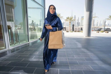 Women's fashion and accessories, a Saudi woman holding paper bags, taking advantage of seasonal offers and discounts and White Friday, an Arab Gulf woman, an Emirati woman wearing an abaya and hijab, shopping in malls with a mobile phone in her hand, the body