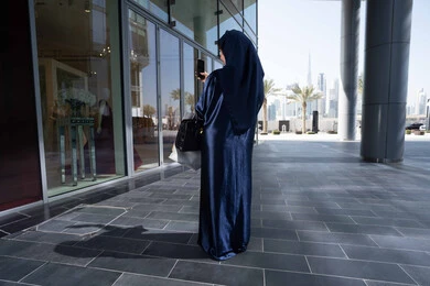 توثيق اللحظات السعيدة، استخدام الأجهزة والتقنيات المتطورة، استغلال الخصومات والتنزيلات، إمرأة سعودية تقوم بالتسوق وشراء المستلزمات والهدايا،صورة من الخلف سيدة عربية خليجية اماراتية ترتدي الحجاب والعباءة تمسك بيدها الهاتف المحمول وأكياس التسوق