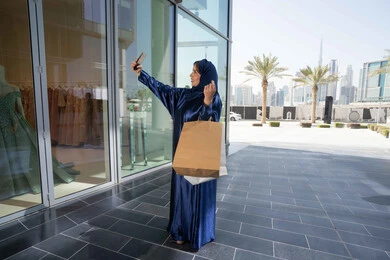 Taking advantage of discounts and sales, using technology in daily life, a Saudi woman shopping and buying supplies and gifts online, an Emirati Gulf Arab woman wearing a hijab and abaya holding a mobile phone and shopping bags, taking souvenir selfies