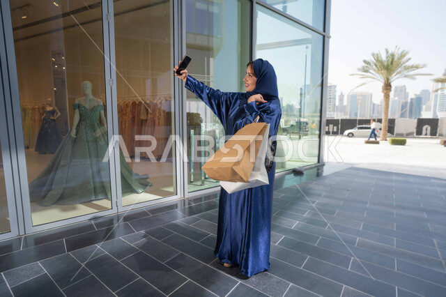 Buying gifts and necessities, taking advantage of seasonal offers and discounts, using technology in daily life, a Saudi woman shopping and buying necessities and gifts, an Emirati Gulf Arab woman wearing a hijab and abaya holding a mobile phone and shopping bags, taking souvenir selfies