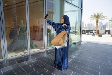 Buying gifts and necessities, taking advantage of seasonal offers and discounts, using technology in daily life, a Saudi woman shopping and buying necessities and gifts, an Emirati Gulf Arab woman wearing a hijab and abaya holding a mobile phone and shopping bags, taking souvenir selfies