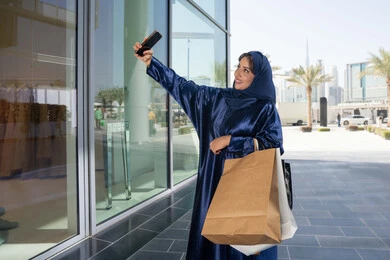التقاط صور السيلفي التذكارية، الاستفادة من العروض الموسمية والتخفيضات، استخدام التكنولوجيا في الحياة اليومية، إمرأة سعودية تقوم بالتسوق وشراء المستلزمات والهدايا عبر الانترنت،سيدة عربية خليجية اماراتية ترتدي الحجاب والعباءة تمسك بيدها الهاتف المحمول وأكياس التسوق