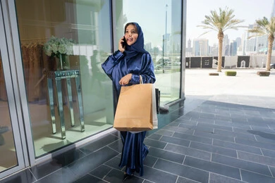 Using technology in daily life, expressions of pleasure and happiness, a Saudi woman shopping and buying supplies and gifts online, an Arab Gulf Emirati woman wearing a hijab and abaya holding a mobile phone and shopping bags in her hand