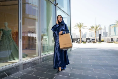 تعابير السرور والسعادة، استخدام التكنولوجيا في الحياة اليومية، إمرأة سعودية تقوم بالتسوق وشراء المستلزمات والهدايا عبر الانترنت،سيدة عربية خليجية اماراتية ترتدي الحجاب والعباءة تمسك بيدها الهاتف المحمول وأكياس التسوق