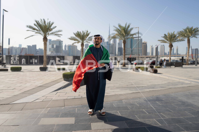 ذكرى يوم الاتحاد والاستقلال الإماراتي 2 ديسمبر، ايماءات الفخر و الإعجاب، صورة من الامام لرجل عربي خليجي اماراتي يرتدي الكندورة و الغترة يضع علم الإمارات على كتفية في النهار، التجول في الشوارع، الجسم بالطول الكامل