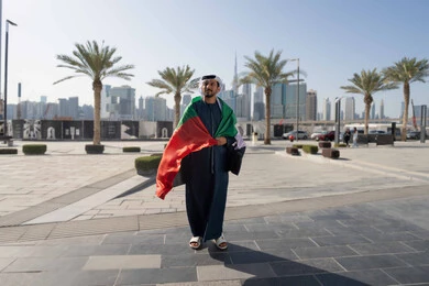 UAE Union and Independence Day, December 2, gestures of pride and admiration, front view of an Emirati Gulf Arab man wearing a kandura and ghutra, holding the UAE flag on his shoulders during the day, walking in the streets, full body