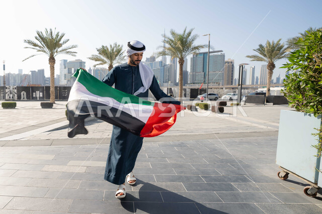 ايماءات السعادة و الفخر و السرور ، إحياء ذكرى استقلال البلاد، مفهوم حب الوطن والانتماء إليه،صورة من الخلف لرجل عربي خليجي إماراتي يرتدي الكندورة والغترة يمسك علم الإمارات المتحدة، الاحتفال باليوم الوطني 2 ديسمبر، الجسم بالطول الكامل