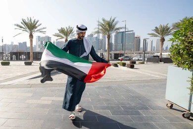 ايماءات السعادة و الفخر و السرور ، إحياء ذكرى استقلال البلاد، مفهوم حب الوطن والانتماء إليه،صورة من الخلف لرجل عربي خليجي إماراتي يرتدي الكندورة والغترة يمسك علم الإمارات المتحدة، الاحتفال باليوم الوطني 2 ديسمبر، الجسم بالطول الكامل