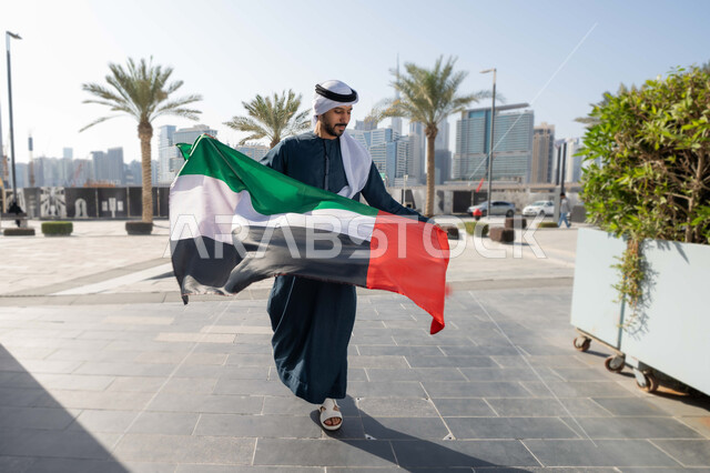 الجسم بالطول الكامل، إحياء ذكرى استقلال البلاد، التجول في الشوارع بايماءات السعادة والفخر والاعتزاز، مفهوم حب الوطن والانتماء إليه،صورة من الخلف لرجل عربي خليجي إماراتي يرتدي الكندورة والغترة يمسك علم الإمارات المتحدة، الاحتفال باليوم الوطني 2 ديسمبر