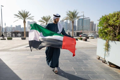 Full body, commemorating the country's independence, walking in the streets with gestures of happiness, pride and honor, the concept of love of the homeland and belonging to it, a picture from the back of an Emirati Gulf Arab man wearing a kandura and ghutra holding the flag of the United Arab Emirates, celebrating the National Day, December 2