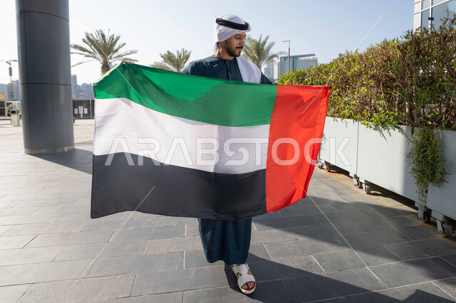 إحياء ذكرى استقلال البلاد، التجول في الشوارع بايماءات السعادة والفخر والاعتزاز، مفهوم حب الوطن والانتماء إليه،صورة من الخلف لرجل عربي خليجي إماراتي يرتدي الكندورة والغترة يمسك علم الإمارات المتحدة، الاحتفال باليوم الوطني 2 ديسمبر، الجسم بالطول الكامل