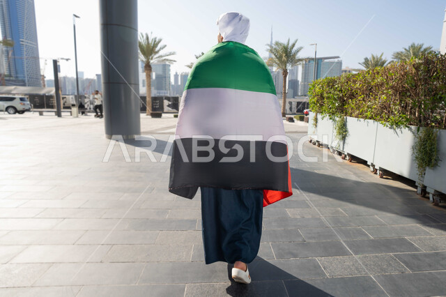ايماءات الفخر و الإعجاب، الاحتفال بذكرى استقلال دولة الإمارات العربية المتحدة، اليوم الوطني الإماراتي 2 ديسمبر، صورة من الخلف لرجل عربي خليجي اماراتي يرتدي الكندورة و الغترة يضع علم الإمارات على كتفية في النهار، التجول في الشوارع