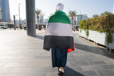 Gestures of pride and admiration, celebrating the anniversary of the independence of the United Arab Emirates, UAE National Day, December 2, a photo from the back of an Emirati Gulf Arab man wearing a kandura and ghutra, placing the UAE flag on his shoulder during the day, walking in the streets
