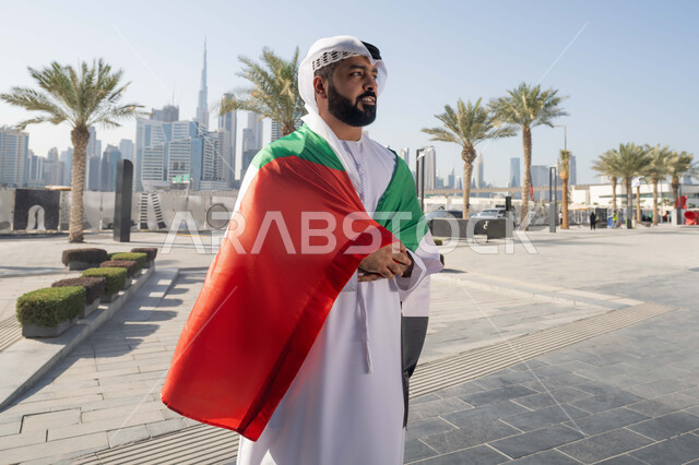 الاحتفال بذكرى استقلال دولة الإمارات العربية المتحدة، اليوم الوطني الإماراتي 2 ديسمبر، رجل عربي خليجي اماراتي يرتدي الكندورة و الغترة يضع علم الإمارات على كتفية في النهار، التجول في الشوارع بايماءات السعادة والفخر والاعتزاز