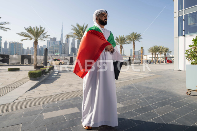اليوم الوطني الإماراتي 2 ديسمبر، الاحتفال بذكرى استقلال دولة الإمارات العربية المتحدة، رجل عربي خليجي اماراتي يرتدي الكندورة و الغترة يضع علم الإمارات على كتفية في النهار، التجول في الشوارع بايماءات السعادة والفخر والاعتزاز،الجسم بالطول الكامل