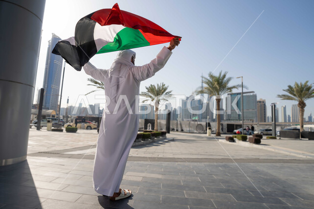 إحياء ذكرى استقلال البلاد، التجول في الشوارع بايماءات السعادة والفخر والاعتزاز، مفهوم حب الوطن والانتماء إليه،صورة من الخلف  لرجل عربي خليجي إماراتي يرتدي الكندورة والغترة يرفع علم الإمارات المتحدة، الاحتفال باليوم الوطني 2 ديسمبر، الجسم بالطول الكامل