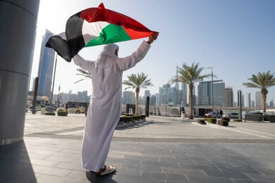 إحياء ذكرى استقلال البلاد، التجول في الشوارع بايماءات السعادة والفخر والاعتزاز، مفهوم حب الوطن والانتماء إليه،صورة من الخلف  لرجل عربي خليجي إماراتي يرتدي الكندورة والغترة يرفع علم الإمارات المتحدة، الاحتفال باليوم الوطني 2 ديسمبر، الجسم بالطول الكامل