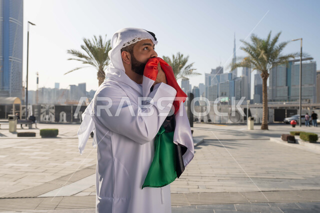 Walking outdoors, UAE National Day identity December 2, UAE National Day 1971, an Emirati Gulf Arab man wearing the traditional ghutra kandara kissing the UAE flag during the day, love and pride of the homeland