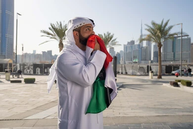 Walking outdoors, UAE National Day identity December 2, UAE National Day 1971, an Emirati Gulf Arab man wearing the traditional ghutra kandara kissing the UAE flag during the day, love and pride of the homeland