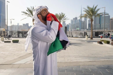 هوية اليوم الوطني الاماراتي 2 ديسمبر، اليوم الوطني الاماراتي 1971، رجل عربي خليجي اماراتي يرتدي الكندرة الغترة التقليدية يقبل علم الامارات العربية المتحدة في النهار، التجول في الهواء الطلق، حب الوطن والاعتزاز به