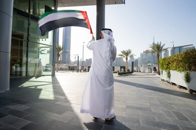 إظهار مشاعر الفخر بالهوية الاماراتية، مفهوم حب الوطن والانتماء إليه، إحياء ذكرى استقلال البلاد،صورة من الخلف لرجل عربي خليجي إماراتي يرتدي الكندورة البيضاء والغترة يرفع علم الإمارات المتحدة، الاحتفال باليوم الوطني 2 ديسمبر، الجسم بالطول الكامل