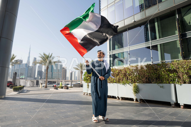 Walking in the streets with gestures of happiness, pride and honor, commemorating the country's independence, the concept of love of the homeland and belonging to it, an Arab Gulf Emirati man wearing a kandura and ghutra raising the flag of the United Arab Emirates, celebrating the National Day, December 2, full body