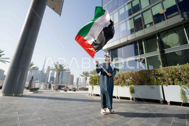 إحياء ذكرى استقلال البلاد، مفهوم حب الوطن والانتماء إليه، رجل عربي خليجي إماراتي يرتدي الكندورة والغترة يرفع علم الإمارات المتحدة، الاحتفال باليوم الوطني 2 ديسمبر، إيماءات الاعتزاز والفخر، صورة الجسم بالطول الكامل