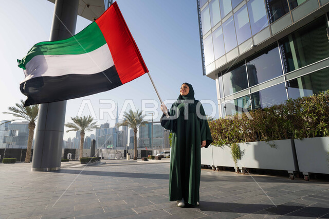 التجول في الشوارع بايماءات السعادة والفخر والاعتزاز، ذكرى تأسيس الاتحاد 2 ديسمبر،استذكار اليوم الوطني الاماراتي 1971، إمراة عربية خليجية إماراتية ترتدي الحجاب والعباءة تمسك بيدها علم الإمارات العربية المتحدة ، يوم العلم 3 نوفمبر، حب الوطن والاعتزاز به
