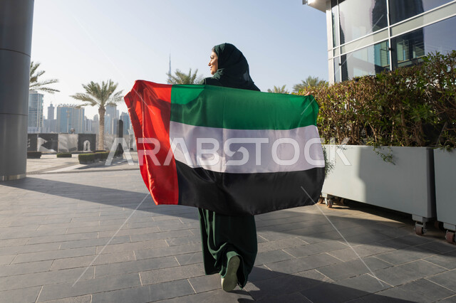 رفع راية الوطن في يوم العلم 3 نوفمبر،  صورة من الخلف لامراة عربية خليجية اماراتية ترتدي العباءة والحجاب تضع علم الإمارات على كتفيها في النهار، التجول في الشوارع بايماءات السعادة والفخر والاعتزاز، مفهوم حب الوطن والانتماء اليه