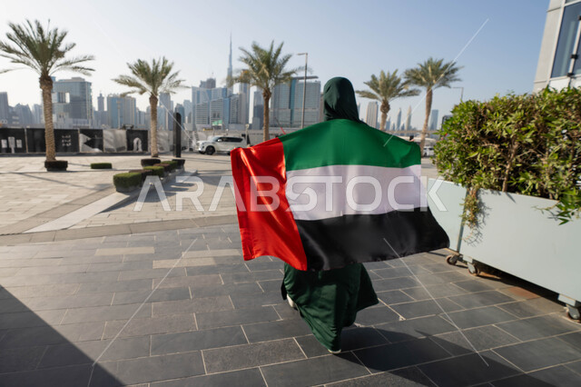 الاحتفال بالاعياد الوطنية للشعوب، لاحتفال بذكرى استقلال دولة الإمارات العربية المتحدة،صورة من الخلف لامراة عربية خليجية اماراتية ترتدي العباءة والحجاب تضع علم الإمارات على كتفيها في النهار، التجول في الشوارع بايماءات السعادة والفخر والاعتزاز، مفهوم حب الوطن والانتماء اليه